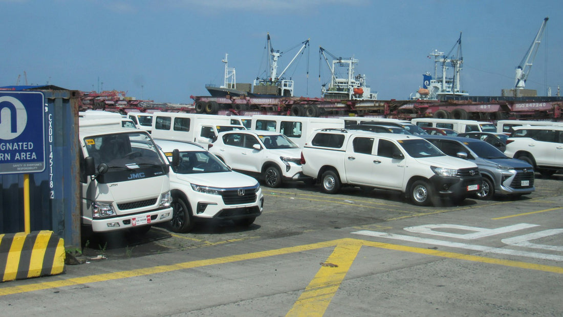 Vehículos blancos estacionados en un puerto junto a buques de carga
