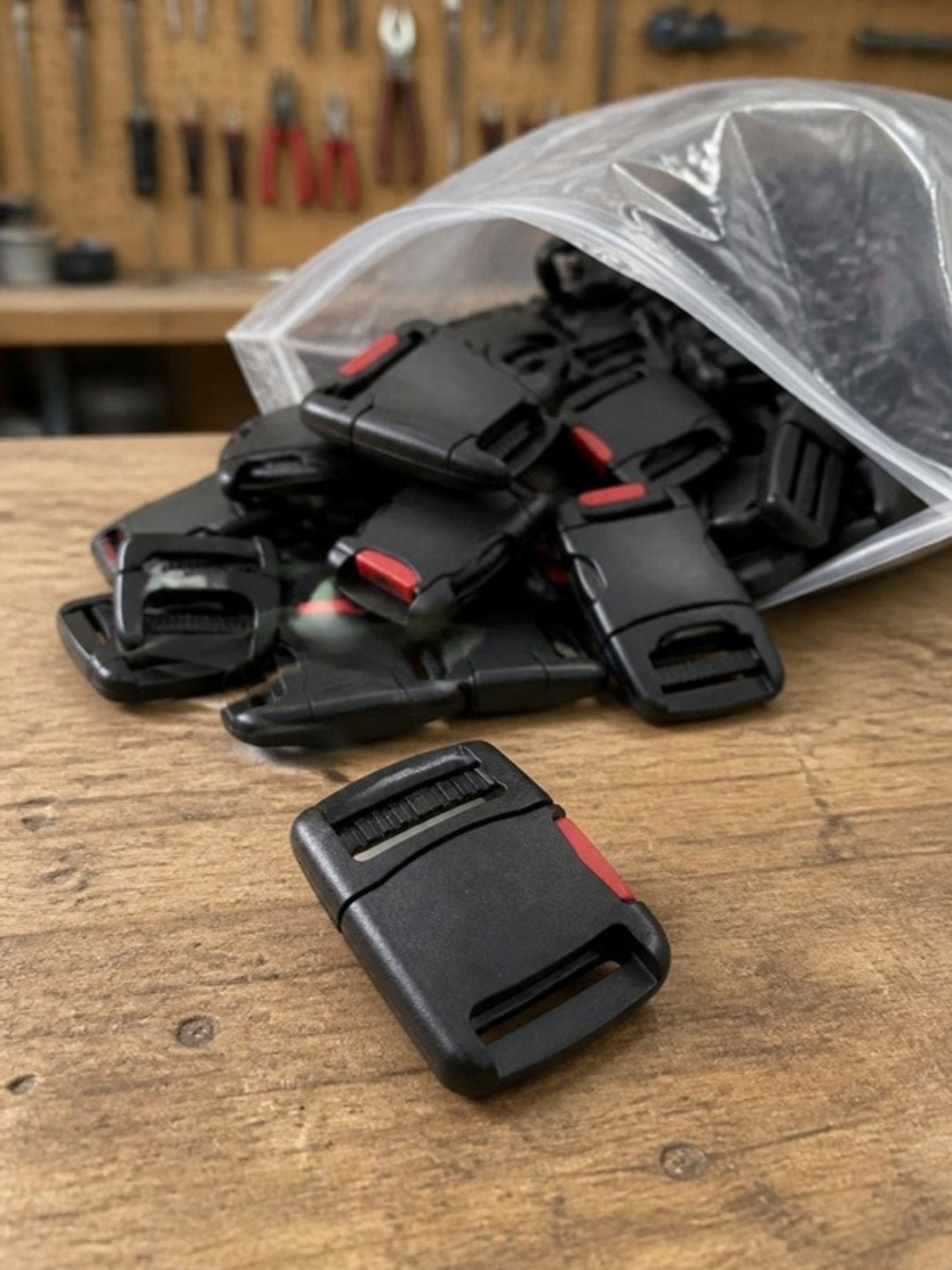 Hebillas laterales de plástico negras con botón rojo saliendo de una bolsa transparente sobre una mesa de taller